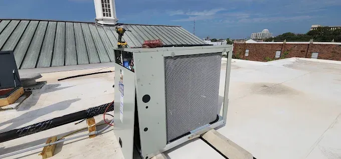 Metal HVAC unit on a white rooftop beside a sloped roof under a clear blue sky