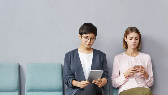 Two women are sitting in a waiting room looking at their phones.