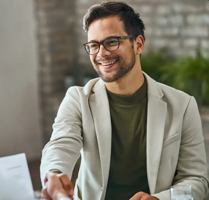 A man wearing glasses is shaking hands with another man while sitting in front of a laptop computer.