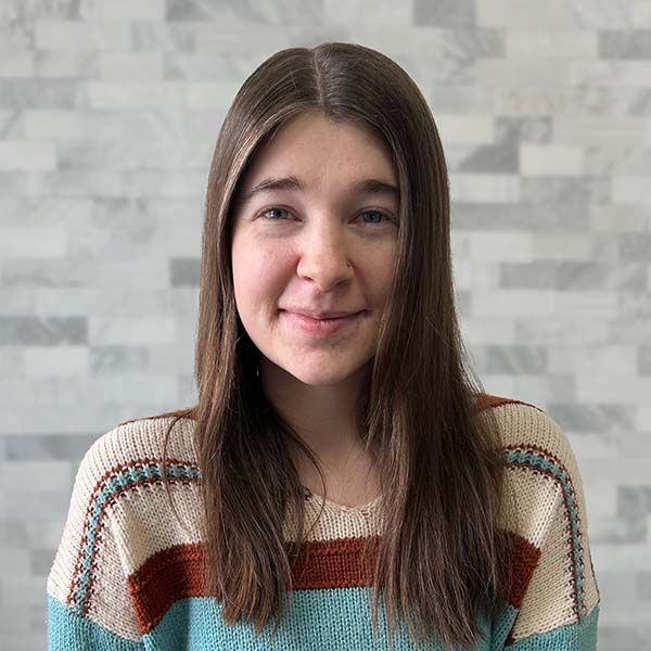 Portrait of a smiling person with long brown hair in a striped sweater against a gray tiled wall