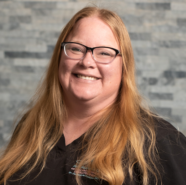 Smiling woman with long red hair and glasses in a black shirt against a gray stone wall.