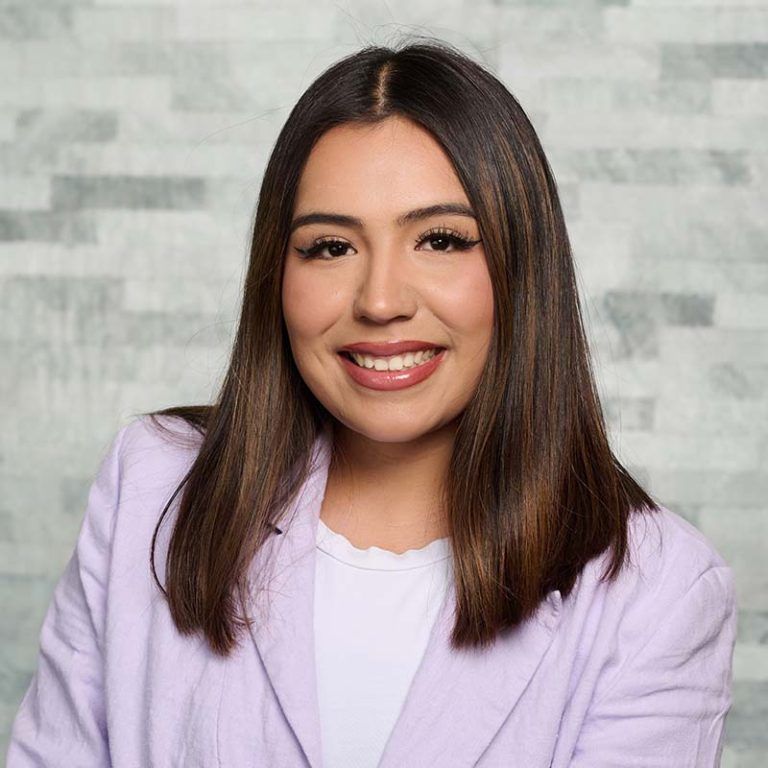Smiling person with shoulder-length dark hair in a light lavender blazer against a gray brick wall