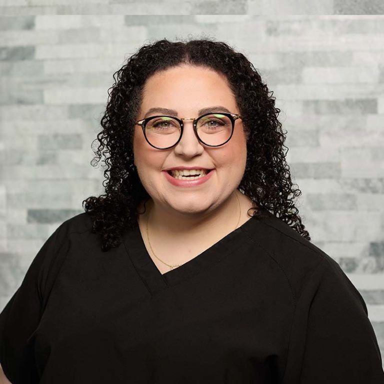 Smiling person with curly hair and glasses, wearing a black top against a light gray brick wall.