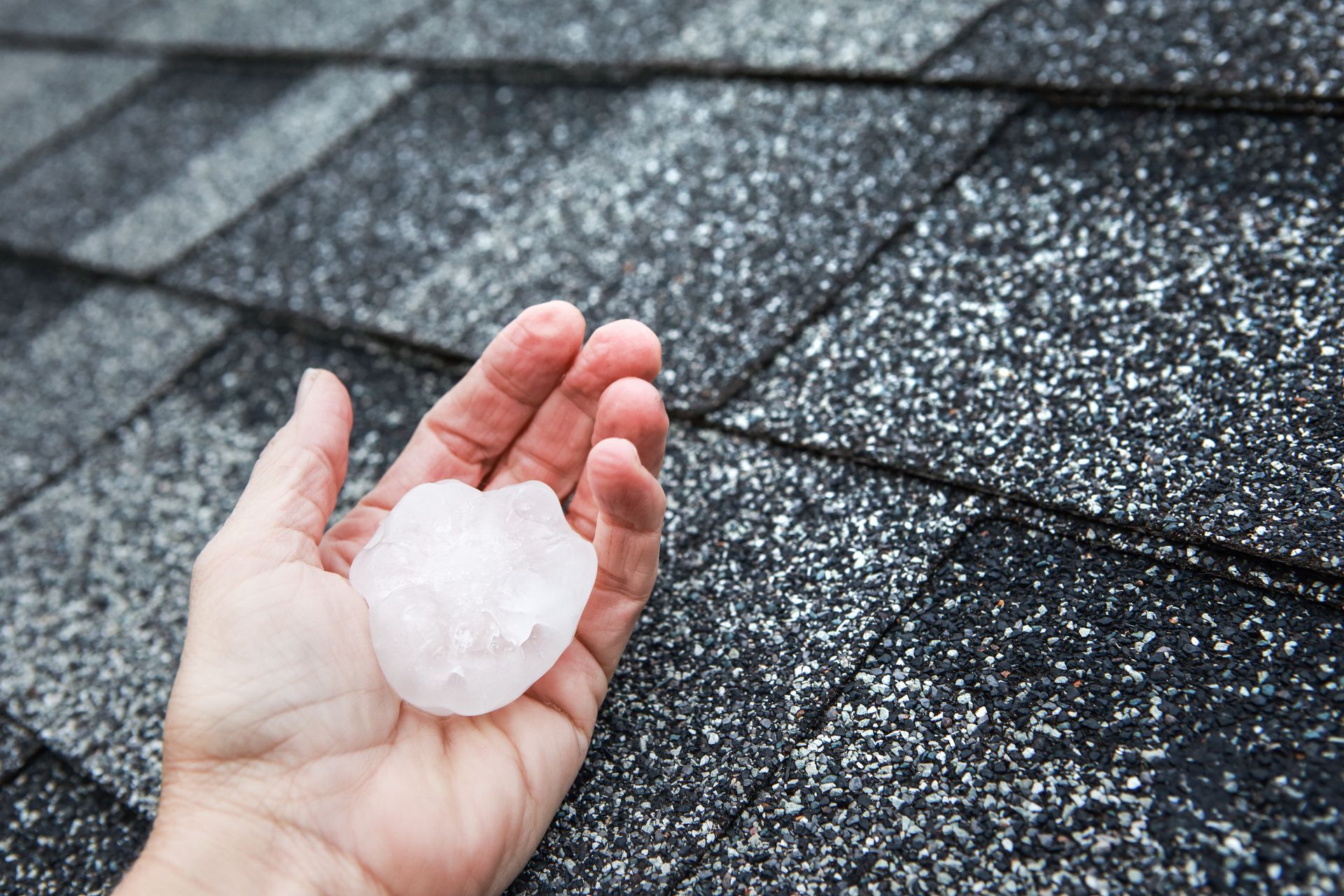 Hand holding large hailstone over asphalt shingles, highlighting need for Storm and Hail Damage Repair services.