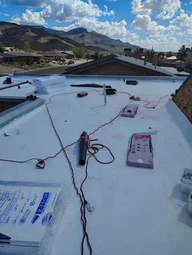 Flat rooftop with white roofing material, tools, and wires, overlooking mountains under a cloudy sky.