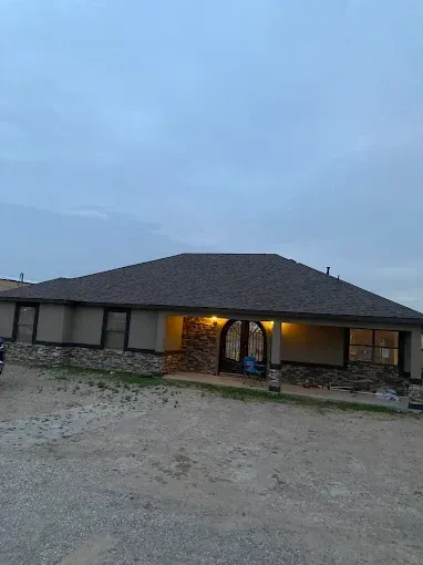 Single-story stone house with lit porch at dusk, set on a gravel driveway under a cloudy sky