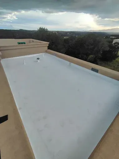Flat white rooftop with beige parapet walls, overlooking trees and hills under a cloudy sky.