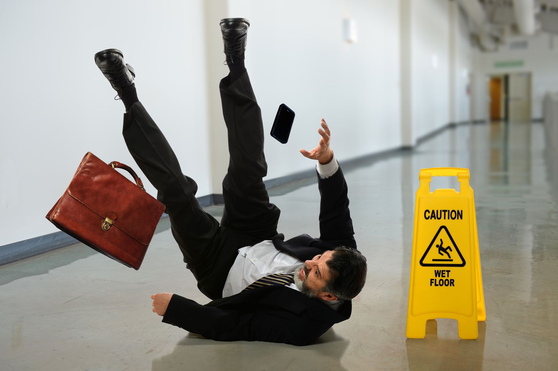 A businessperson slipping on a wet floor in a hallway with a caution sign A businessperson slipping on a wet floor in a hallway with a caution sign