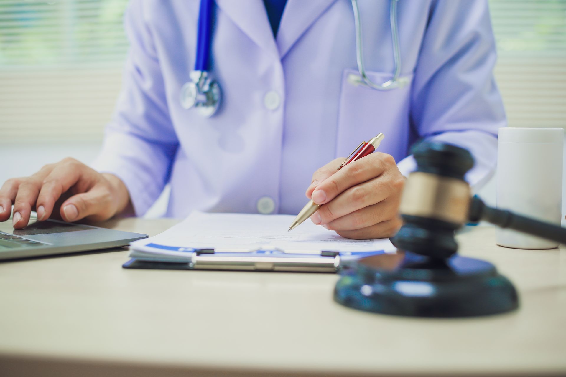 A male doctor signs on a file, with a gavel on a table in front of him.