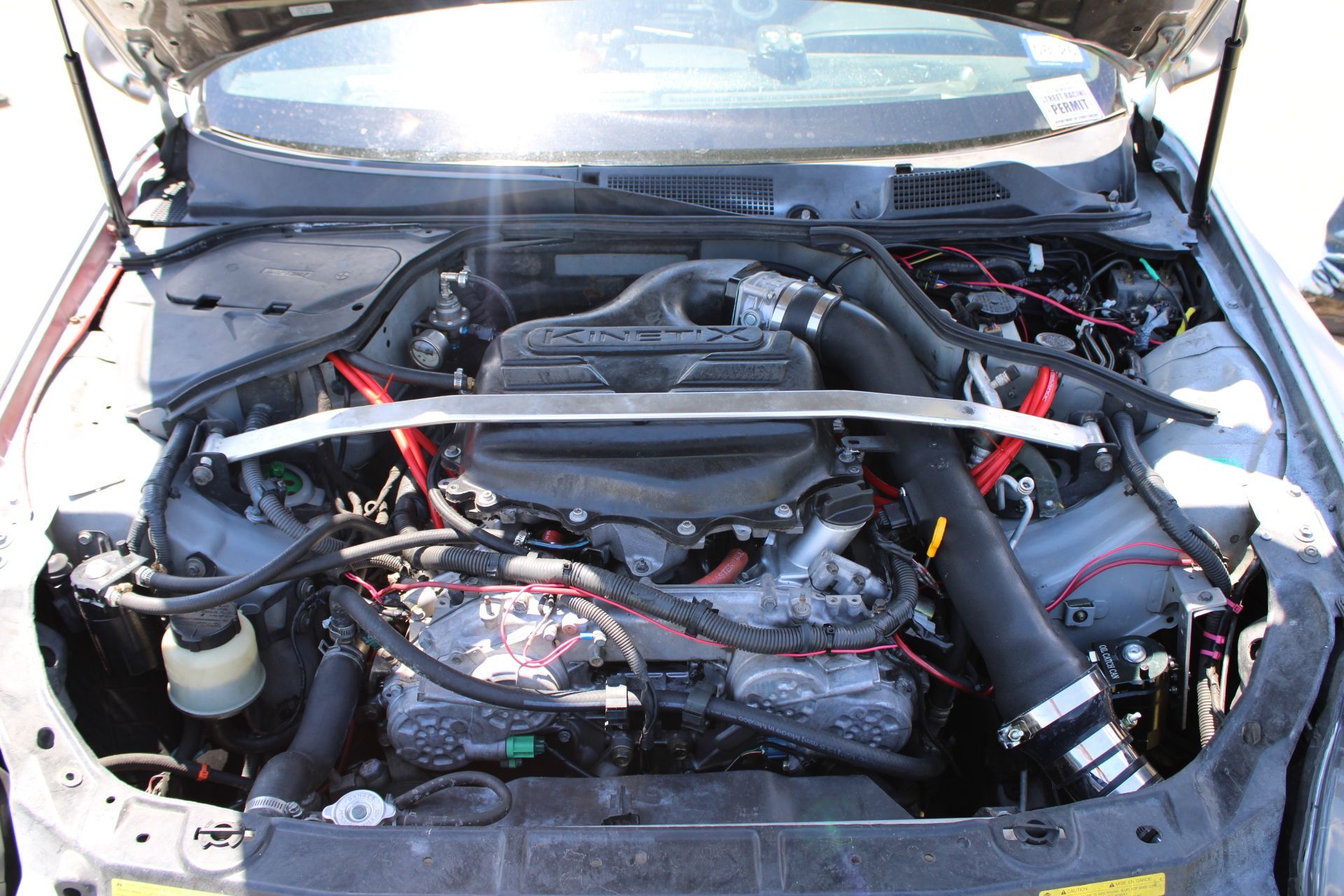 Car engine bay with exposed components, hoses, and a silver strut bar under the open hood