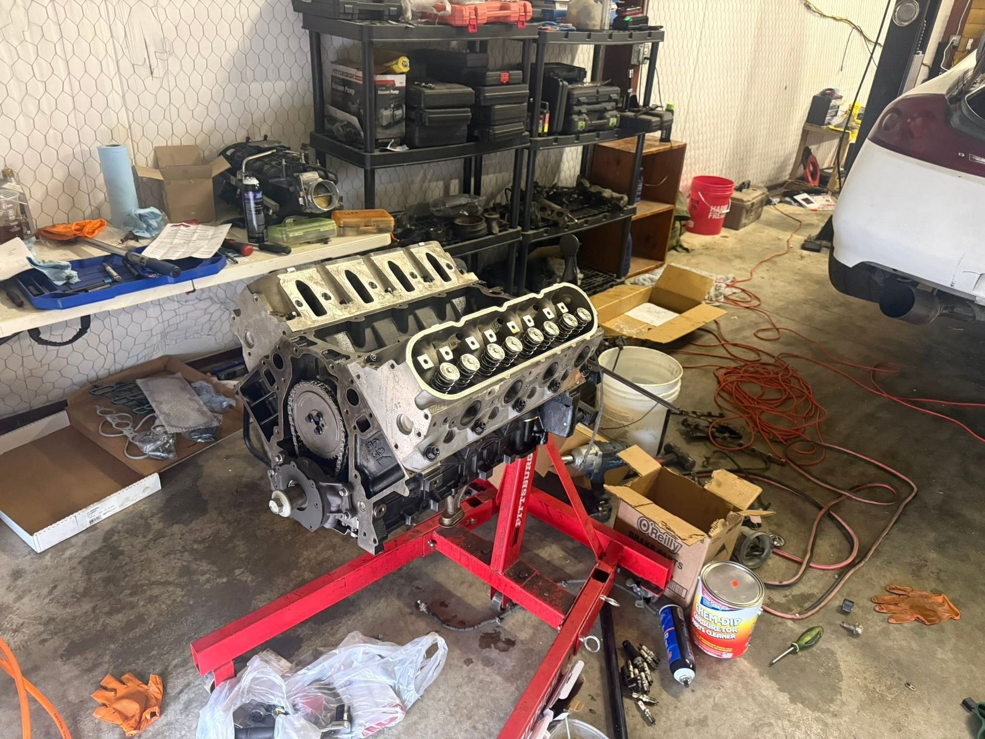 Engine on a red stand in a cluttered garage workshop, with tools and car parts around it.