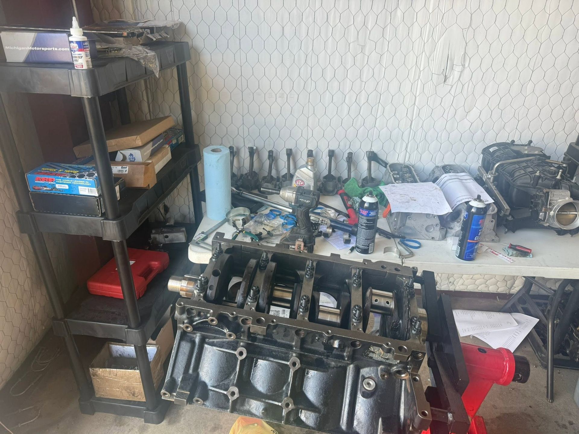 Workshop bench with an engine block, tools, parts, and bottles on shelves in a small garage space