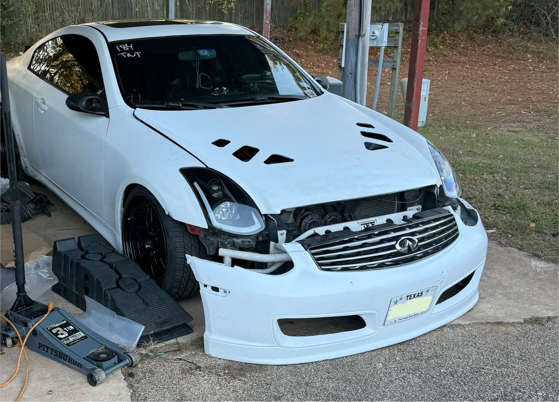 Damaged white car with missing front bumper and hood vents, parked beside repair tools under a carport