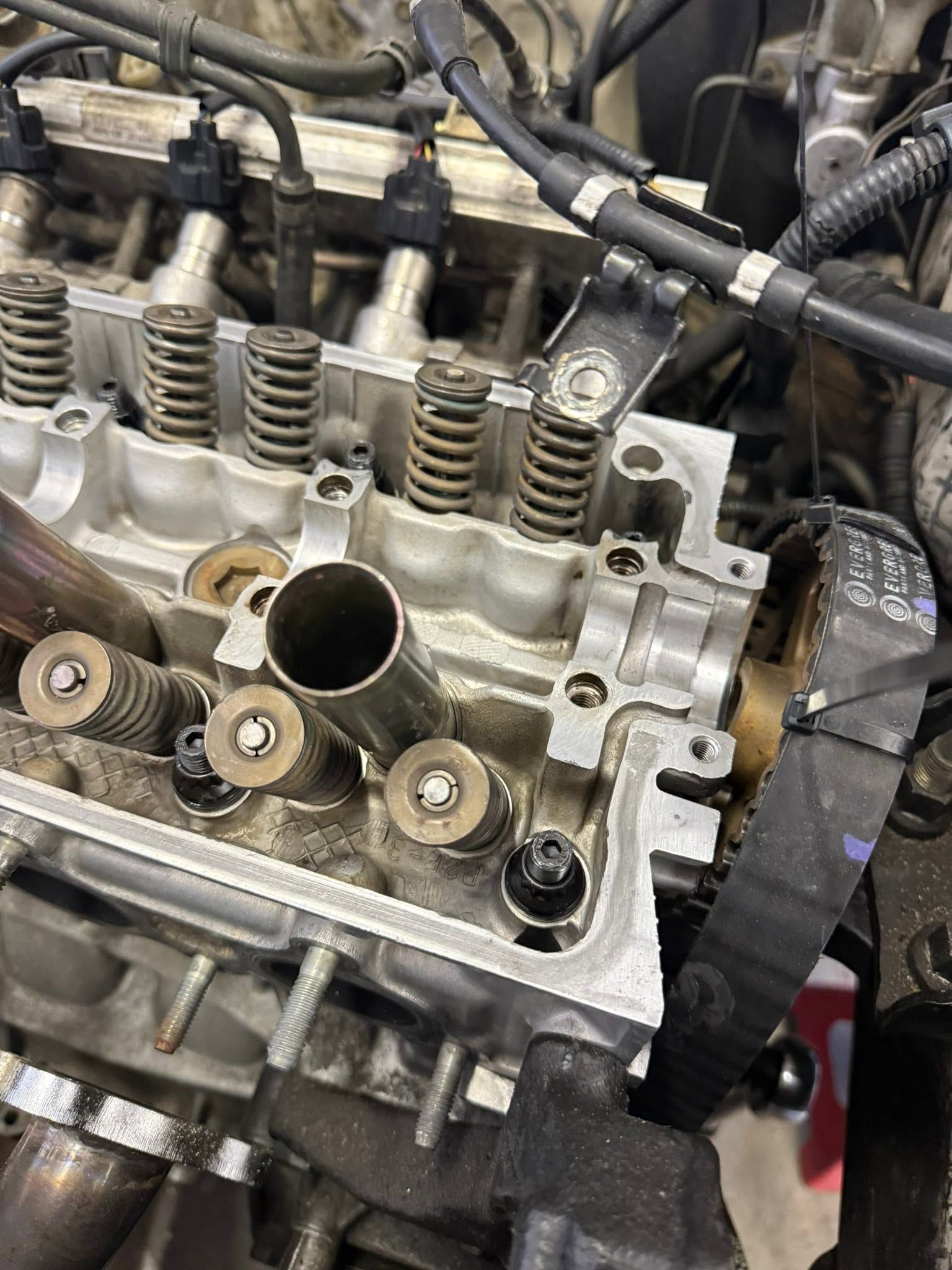 Close-up of an engine cylinder head with exposed valve springs and hoses in a workshop.