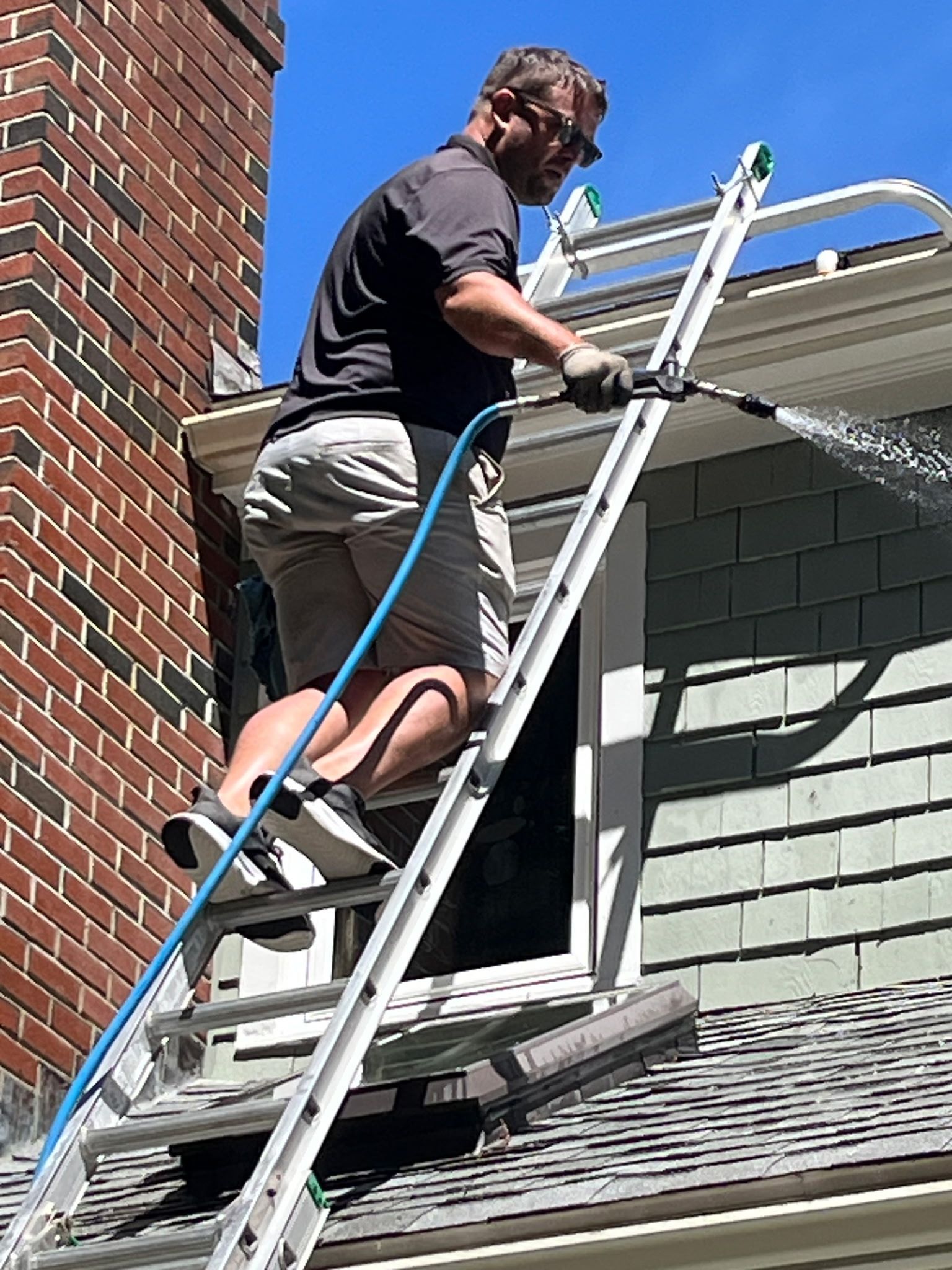 Person in gloves cleaning a gutter filled with leaves. Roof and wall in background.