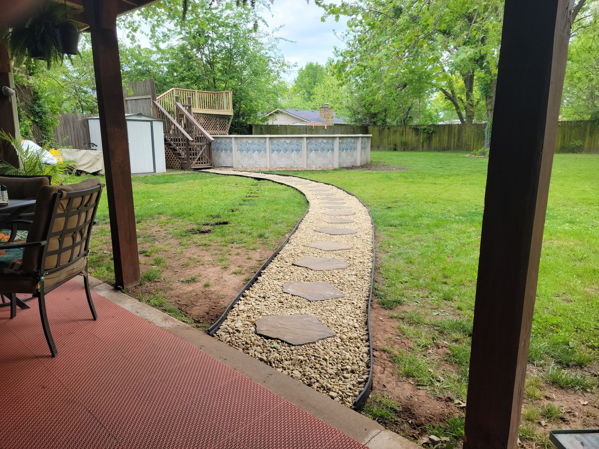 Curved gravel path across a green backyard, viewed from a covered patio with a chair and wooden posts.