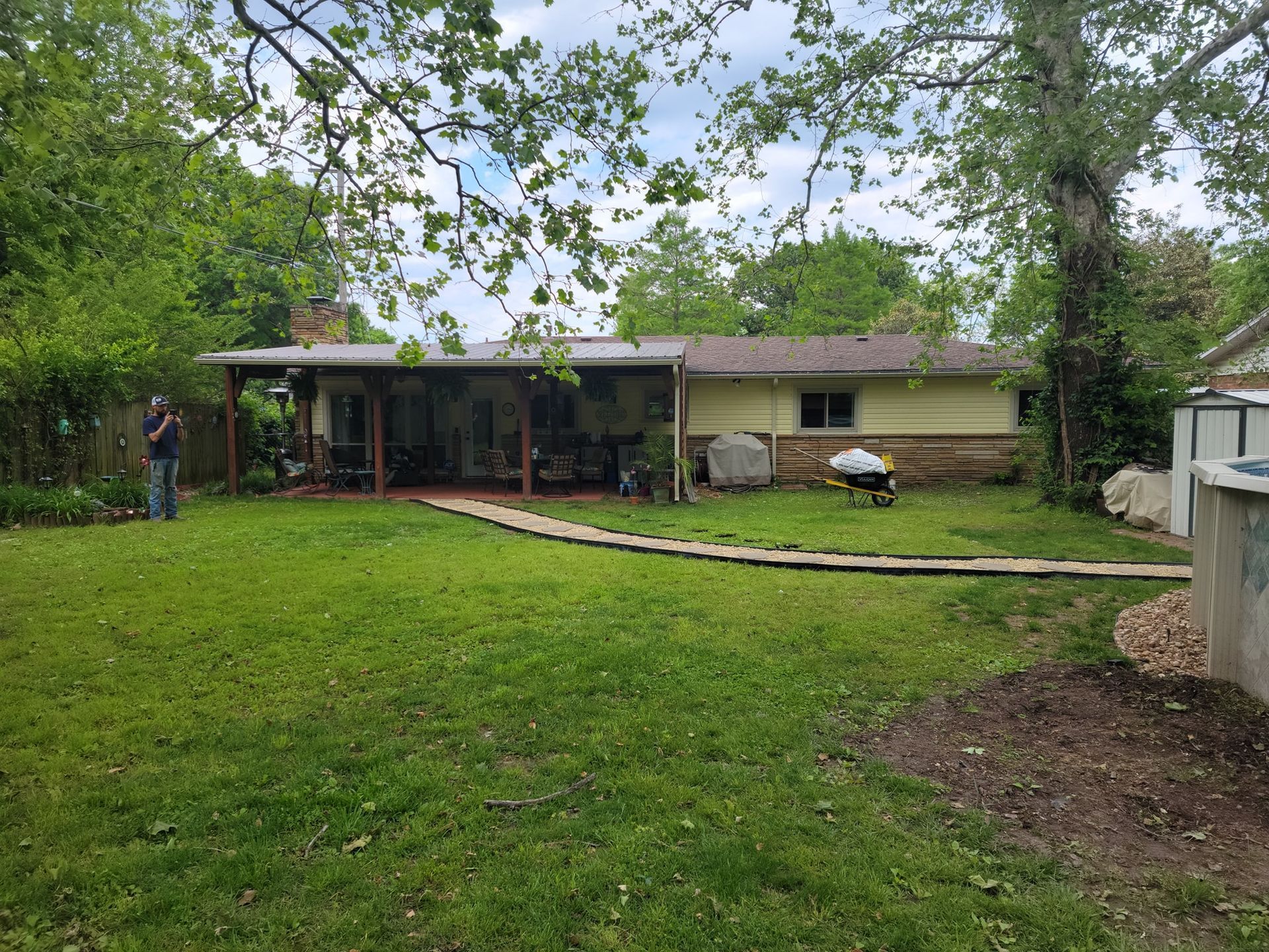 Backyard with a green lawn, patio, and single-story house under trees