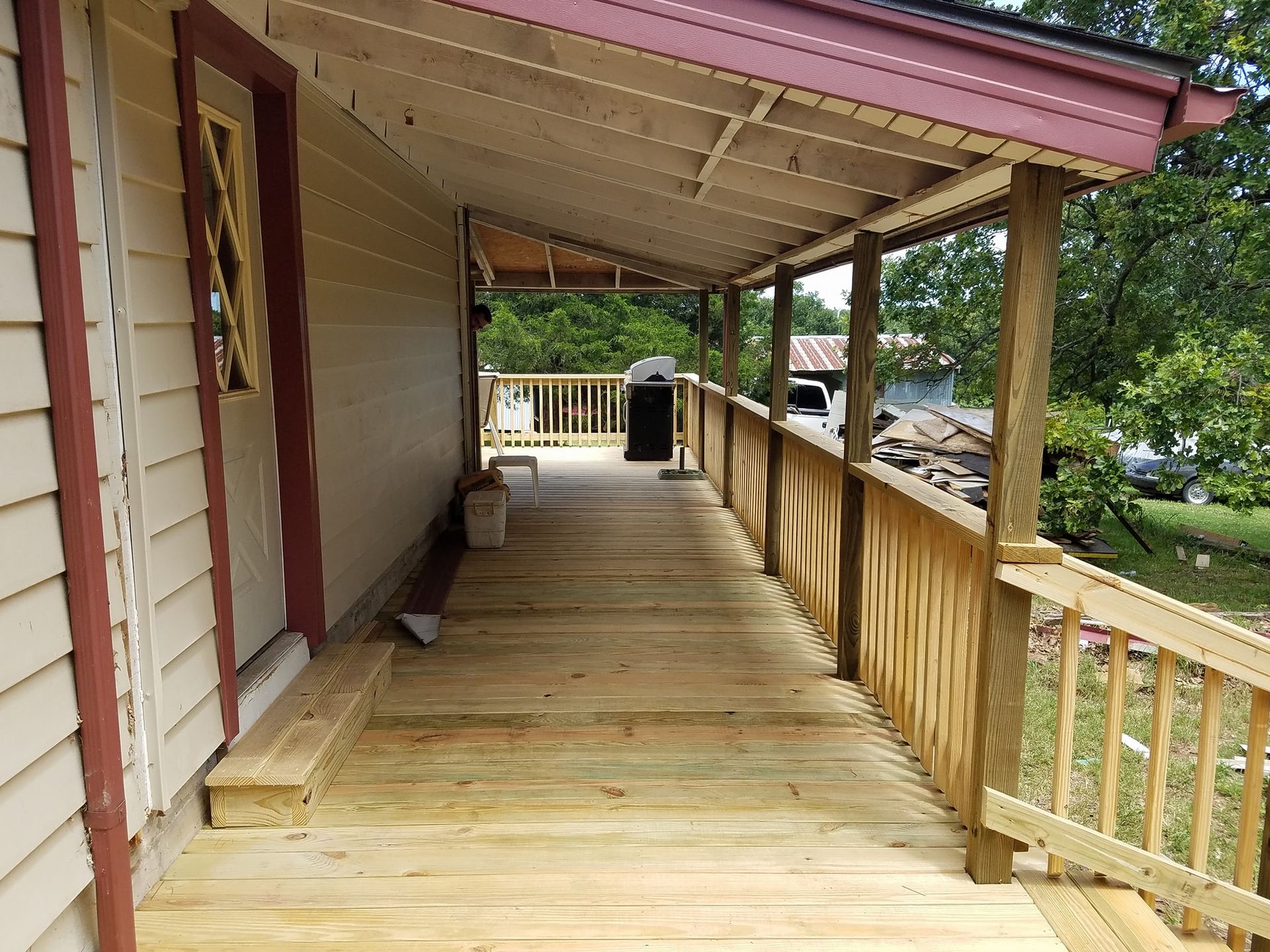 Wooden covered porch with railing, beige siding, and a view of trees and yard.