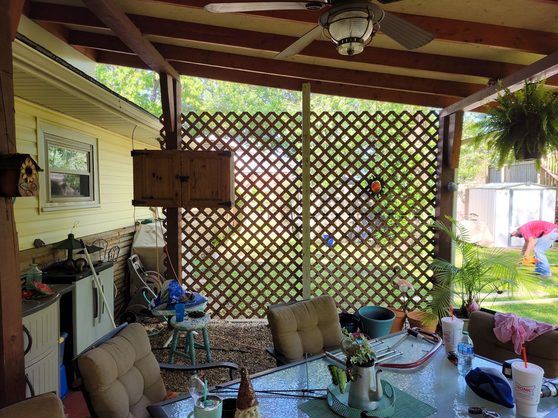 Covered patio with lattice wall, hanging plants, outdoor furniture, and a grill near a grassy yard.