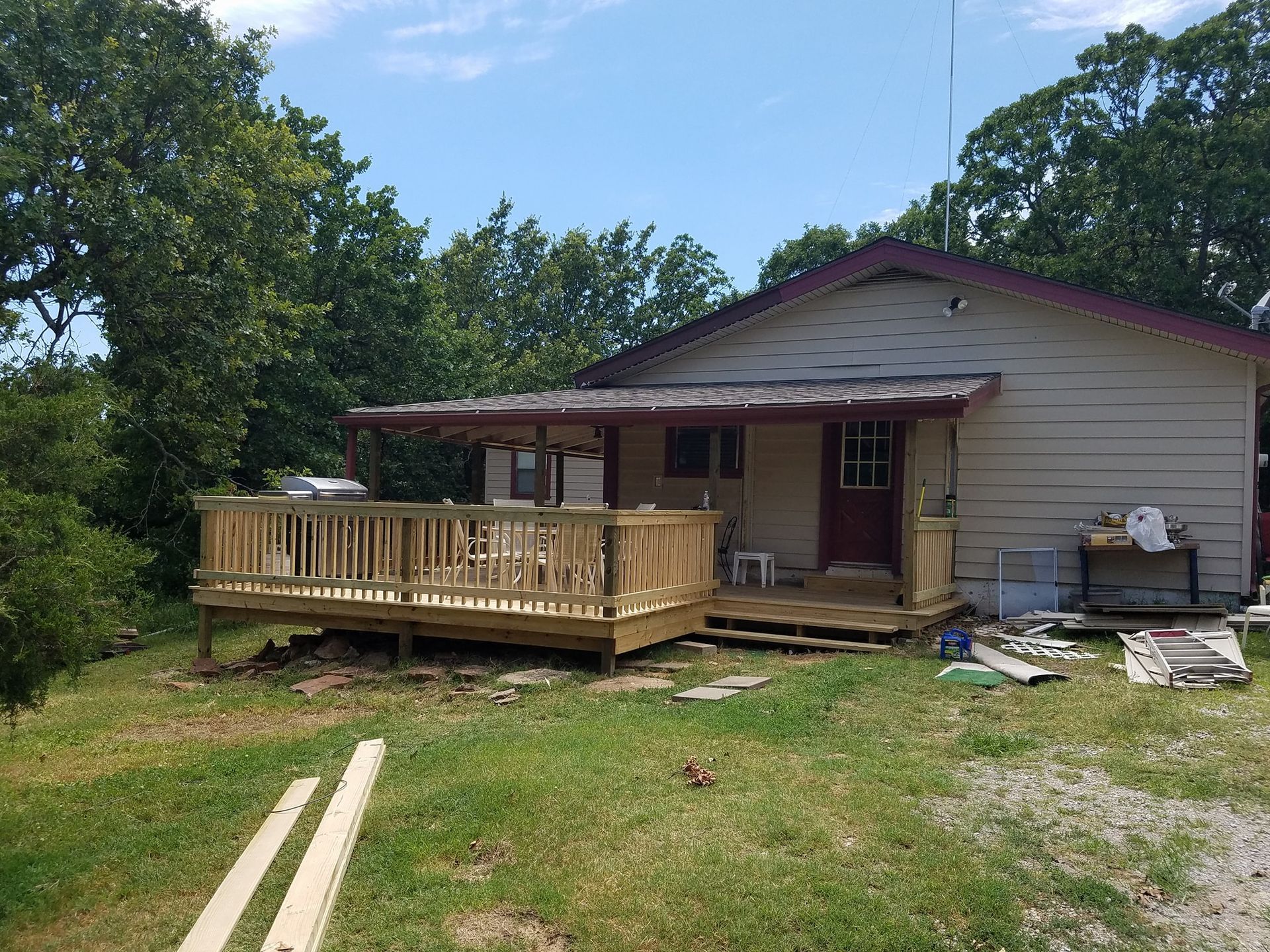 Small house with a wooden front deck in a grassy yard, surrounded by trees and scattered construction materials.