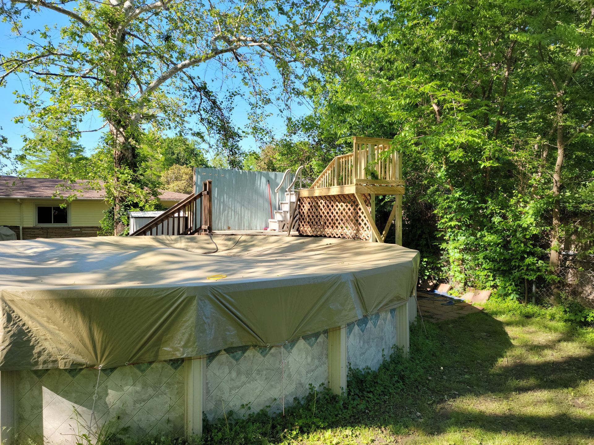 Above-ground pool with tan cover, wooden deck, and trees in a backyard yard