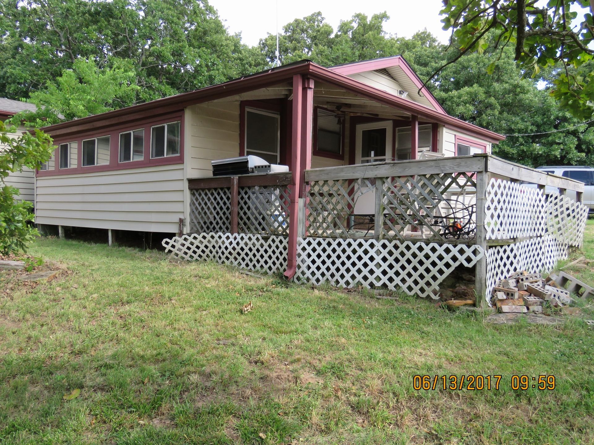 Small beige house with red trim, porch, and lattice railing on a grassy hillside, surrounded by trees.