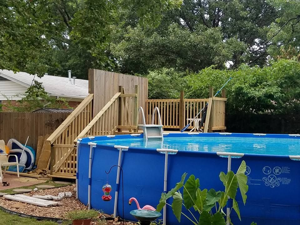 Backyard above-ground pool beside a wooden deck and stairs, with trees and fencing in the background
