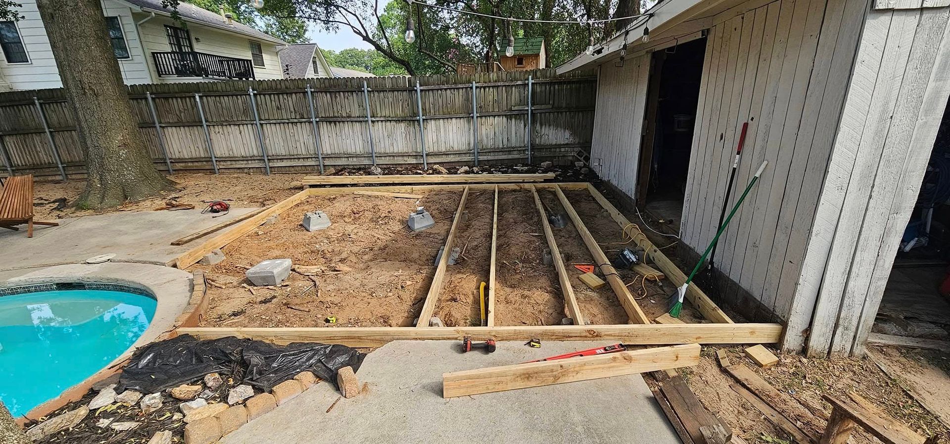 Backyard patio under construction with excavated dirt, wooden forms, and a small pool on the left