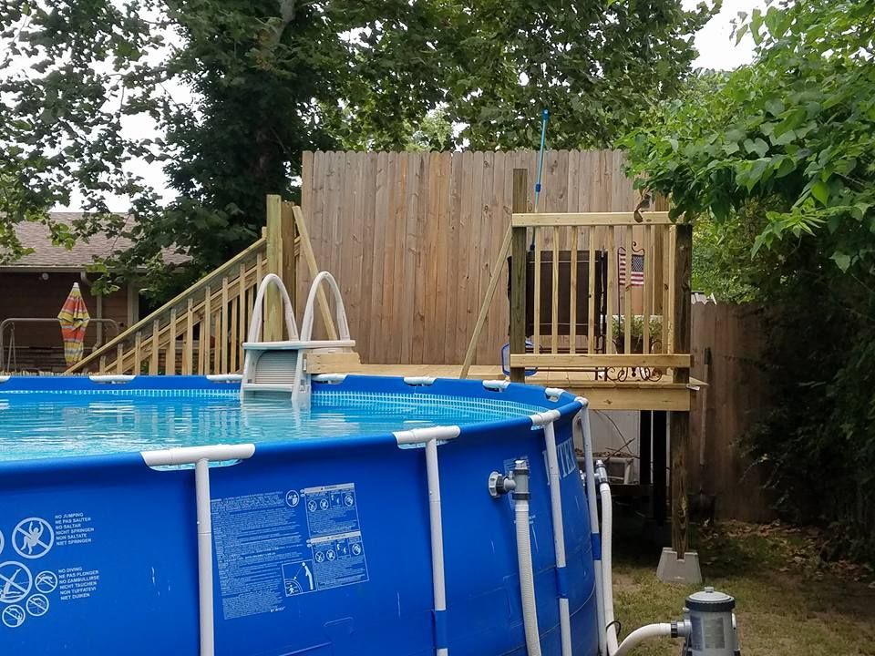 Blue above-ground pool with wooden deck and ladder in a backyard, surrounded by trees and fencing