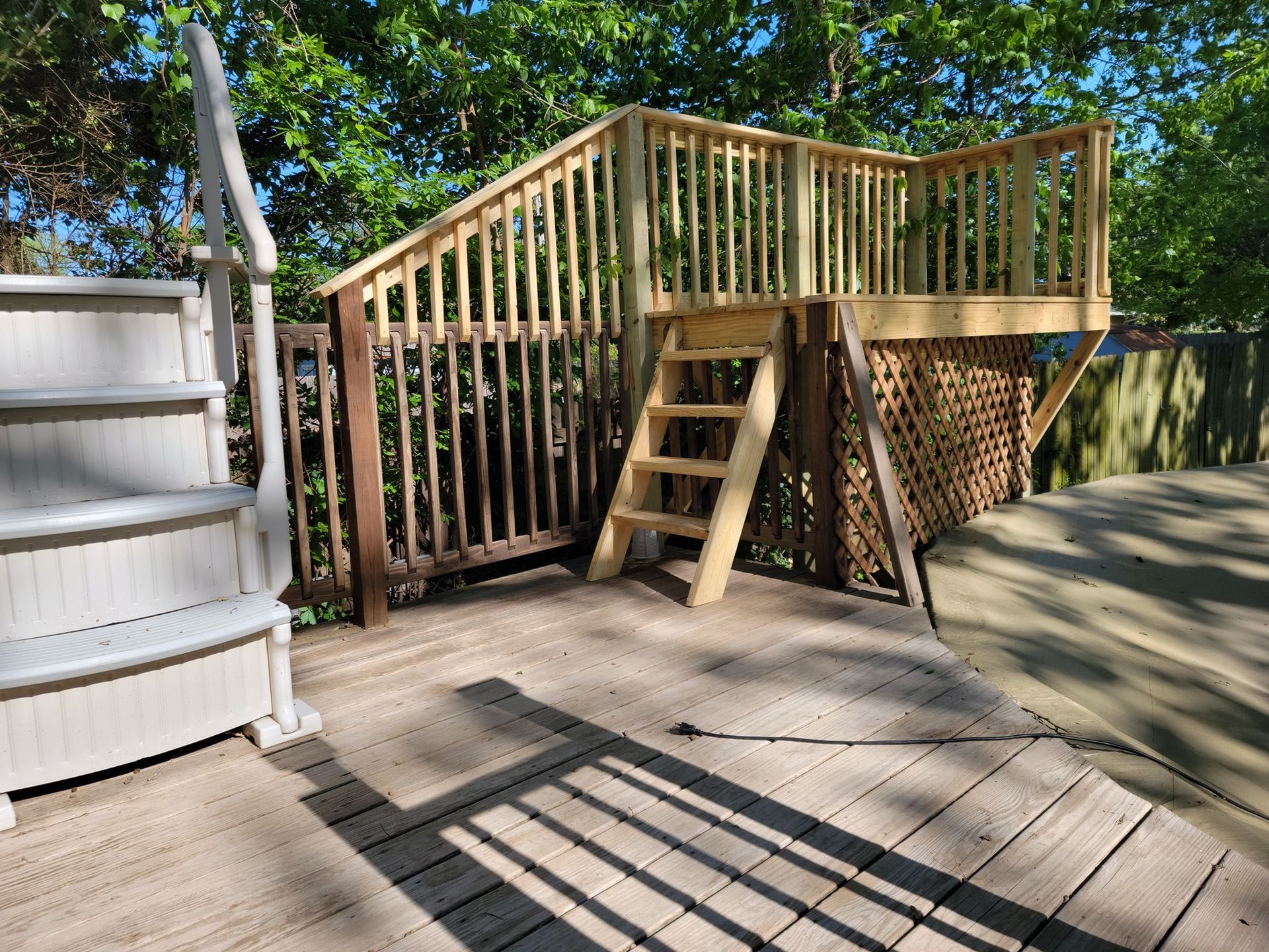 Wooden deck with railing and stairs in a sunny backyard, beside a small shed and trees