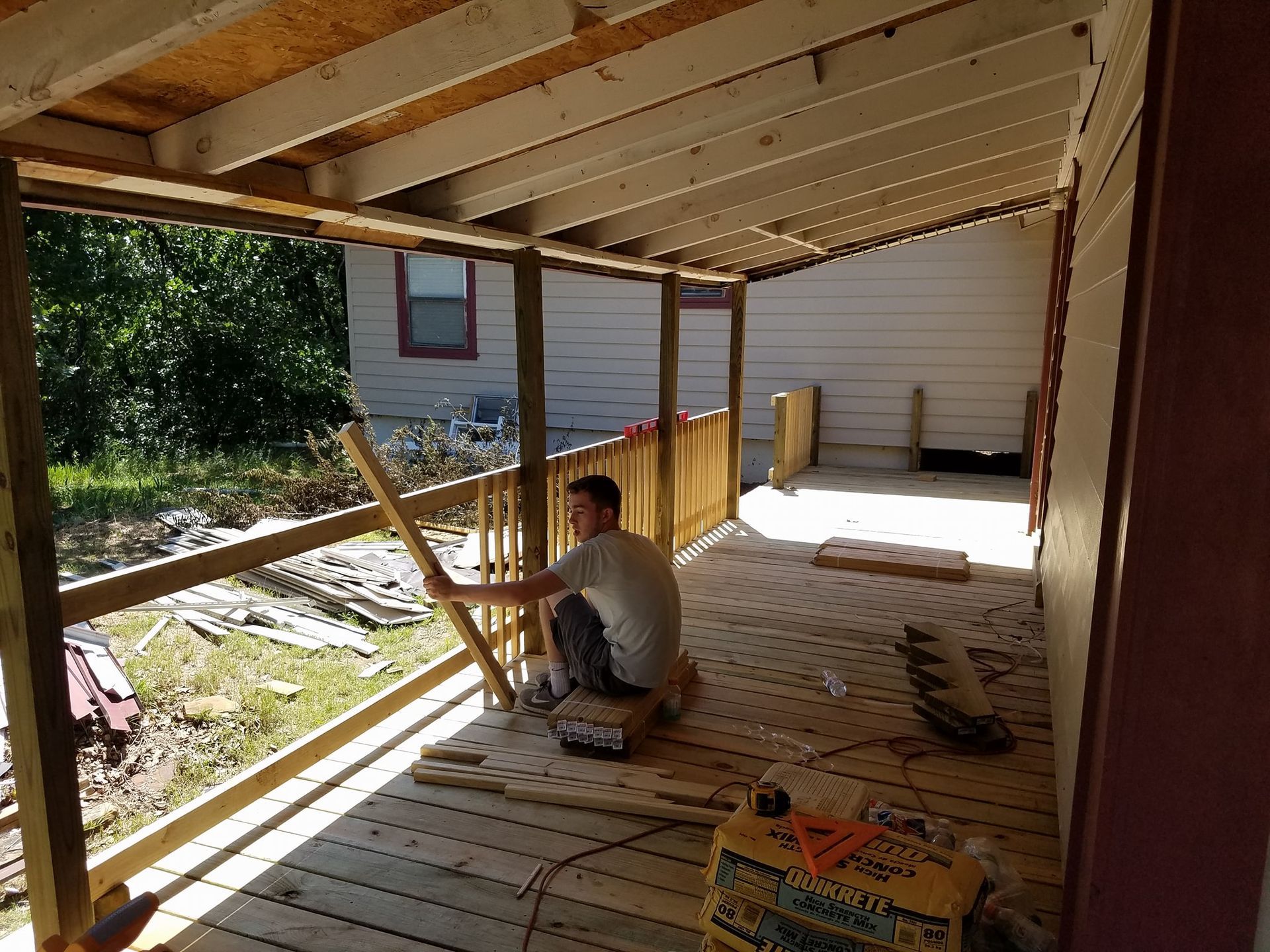 Person working on a wooden deck under construction beside a house, with exposed beams and tools nearby.