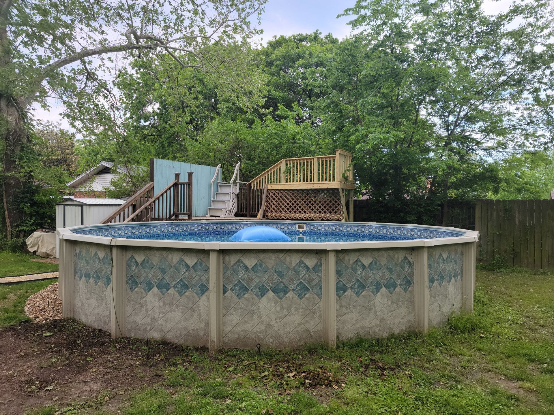Above-ground backyard pool with blue cover, wood deck, and trees surrounding it
