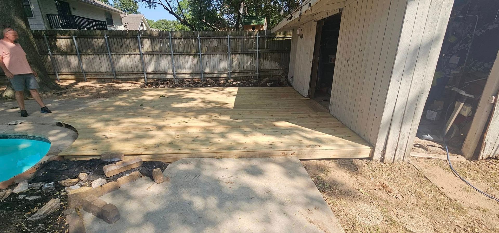 Backyard patio with a person near a small pool, concrete slab, stone wall, and a shed.