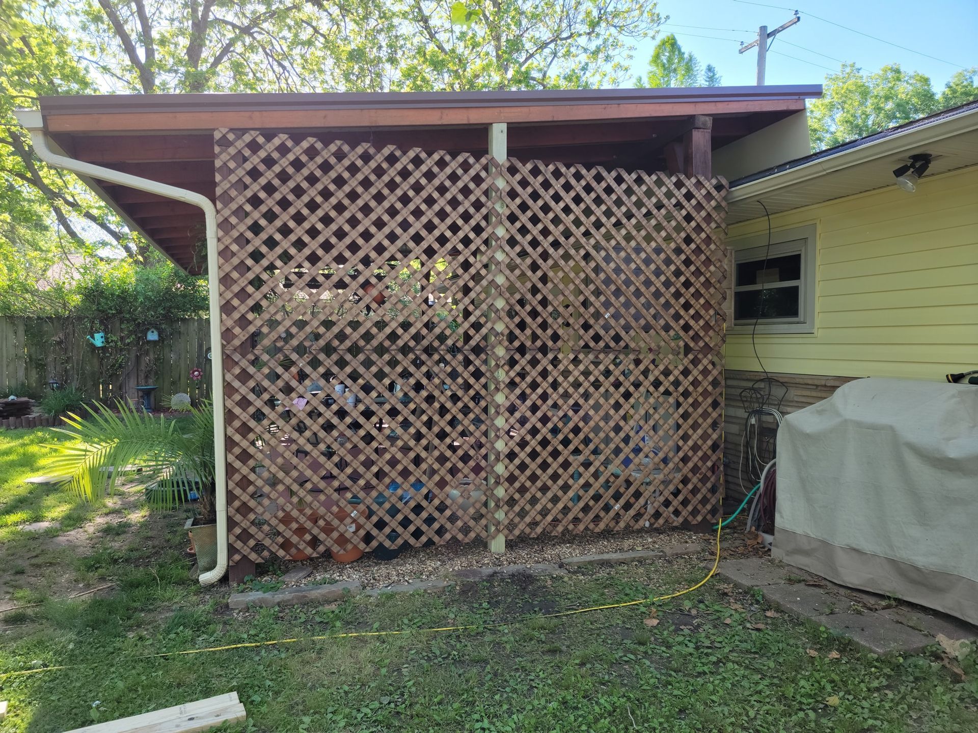Wooden lattice patio enclosure beside a yellow house, with a covered object and yard greenery.