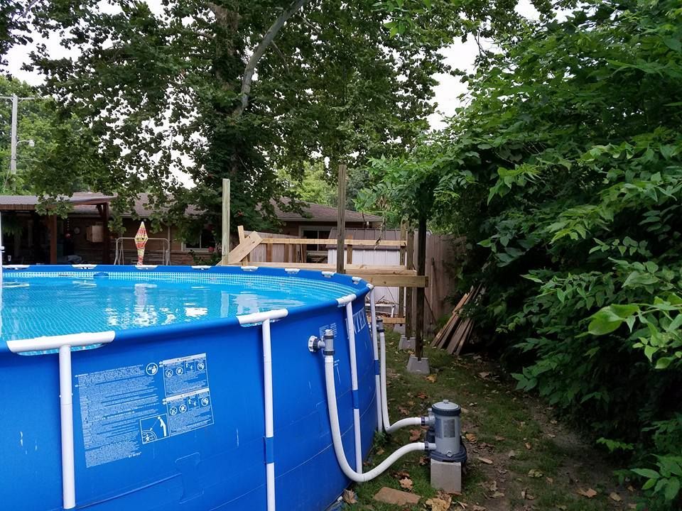 Above-ground blue pool with pump and hose in a leafy backyard with a deck