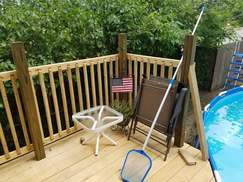 Patio by a pool with a white stool, ladder, broom, and a small American flag on a wooden railing.
