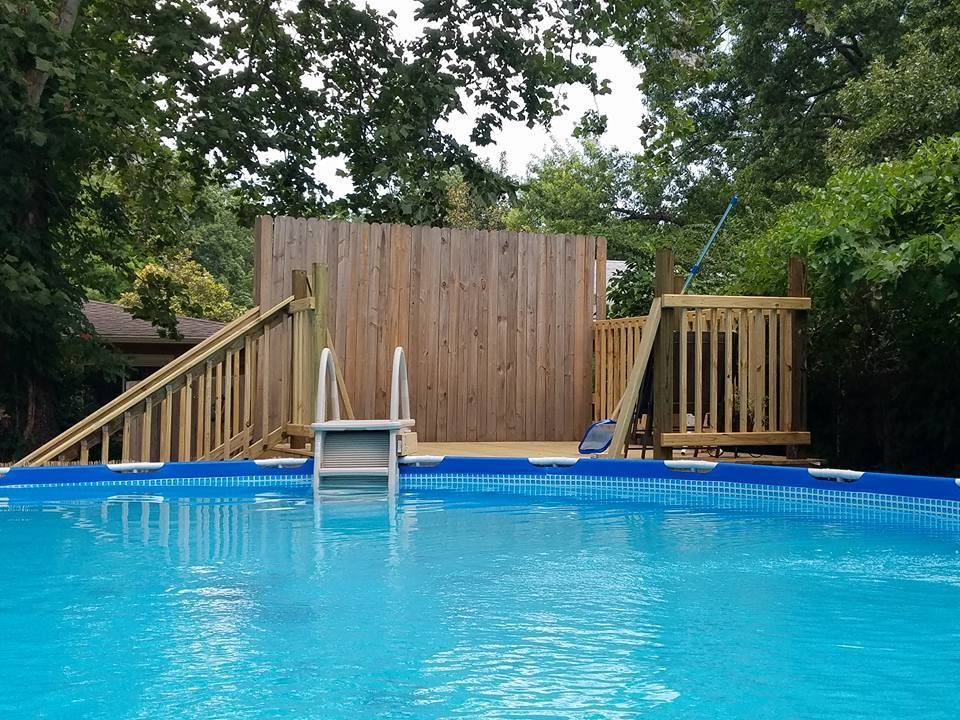 Above-ground pool with wooden deck and ladder, backed by a tall fence and trees