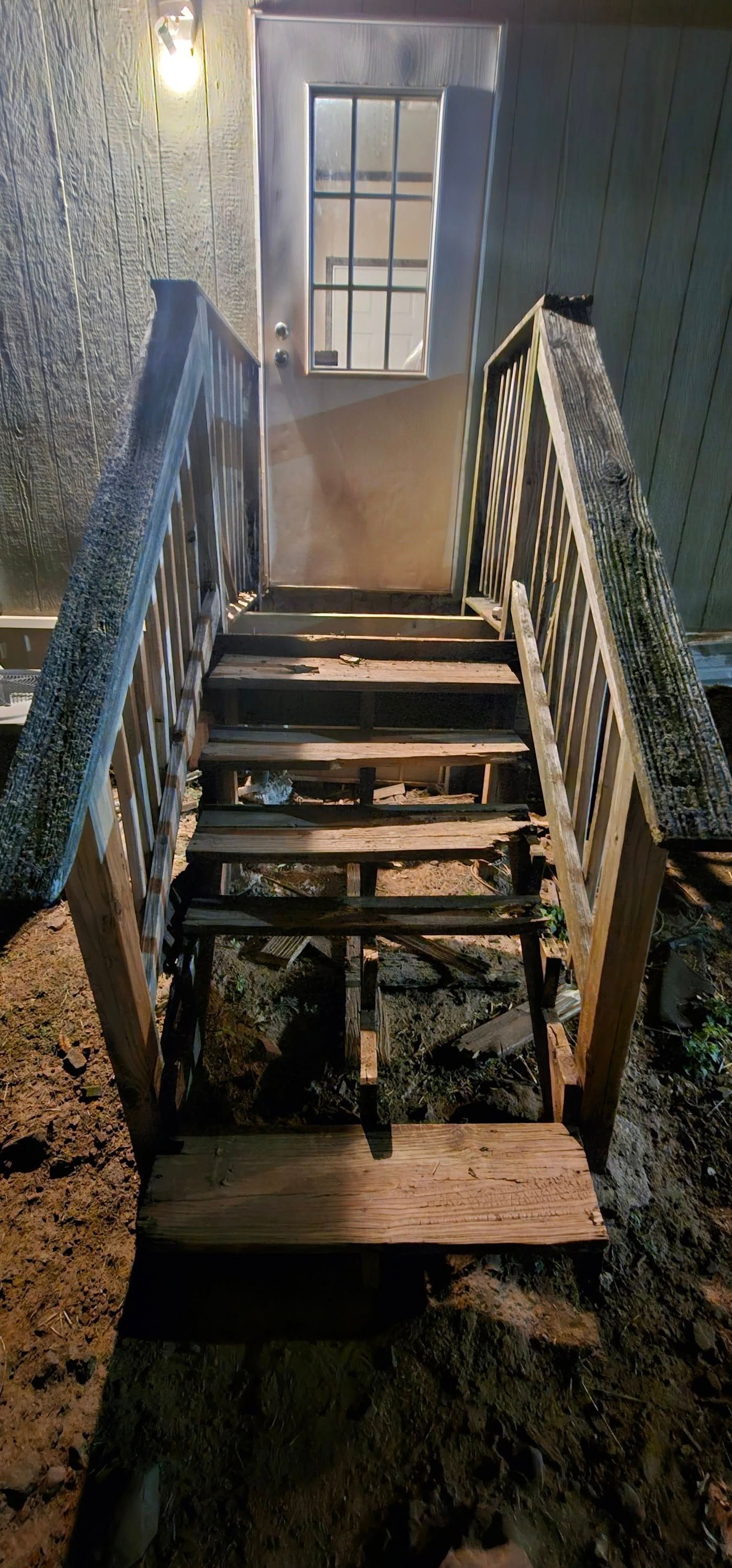 Wooden staircase with railings leading to a lit door at the top on a dim porch at night
