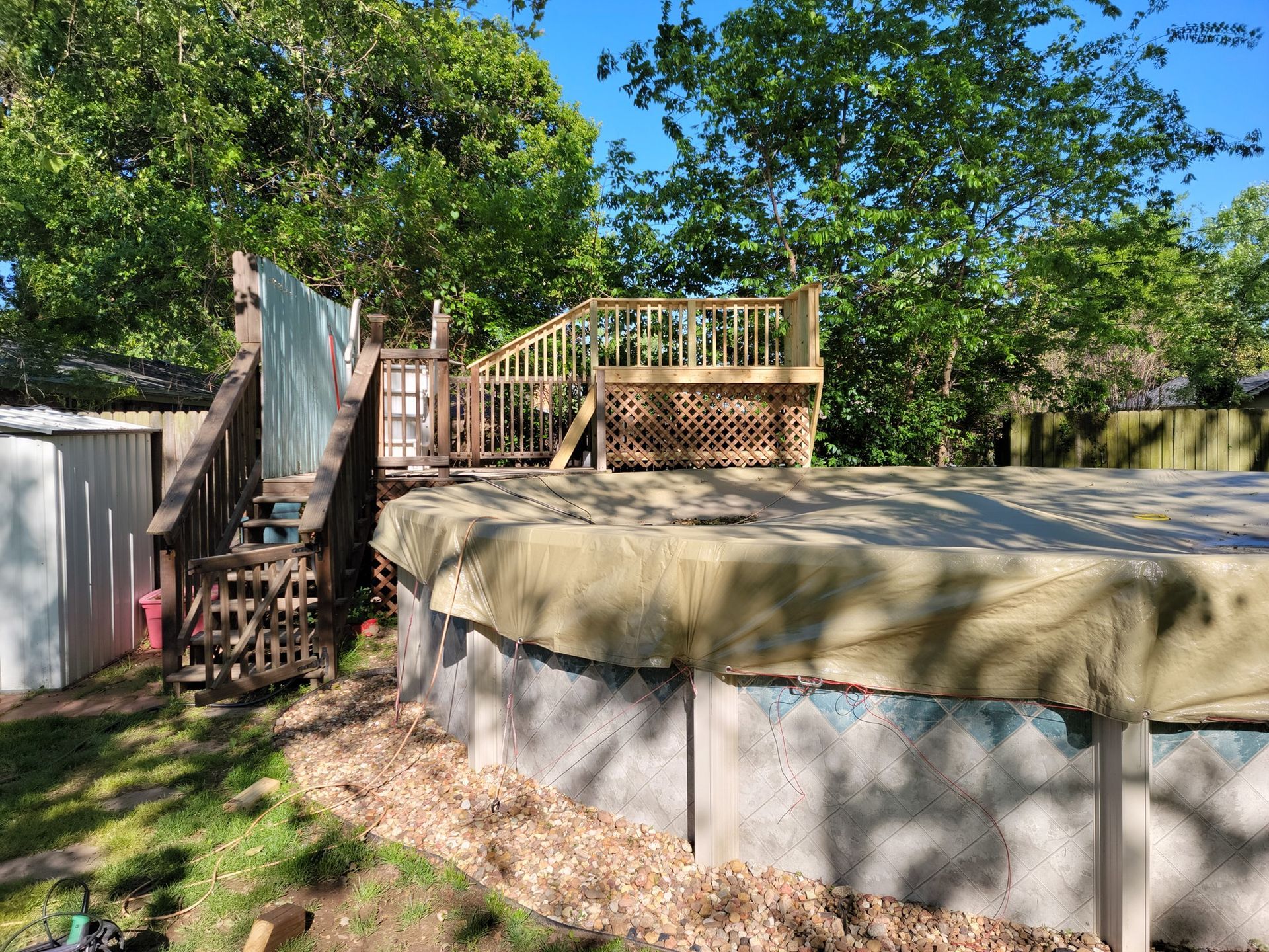 Backyard above-ground pool with wooden deck and steps under trees in sunny yard