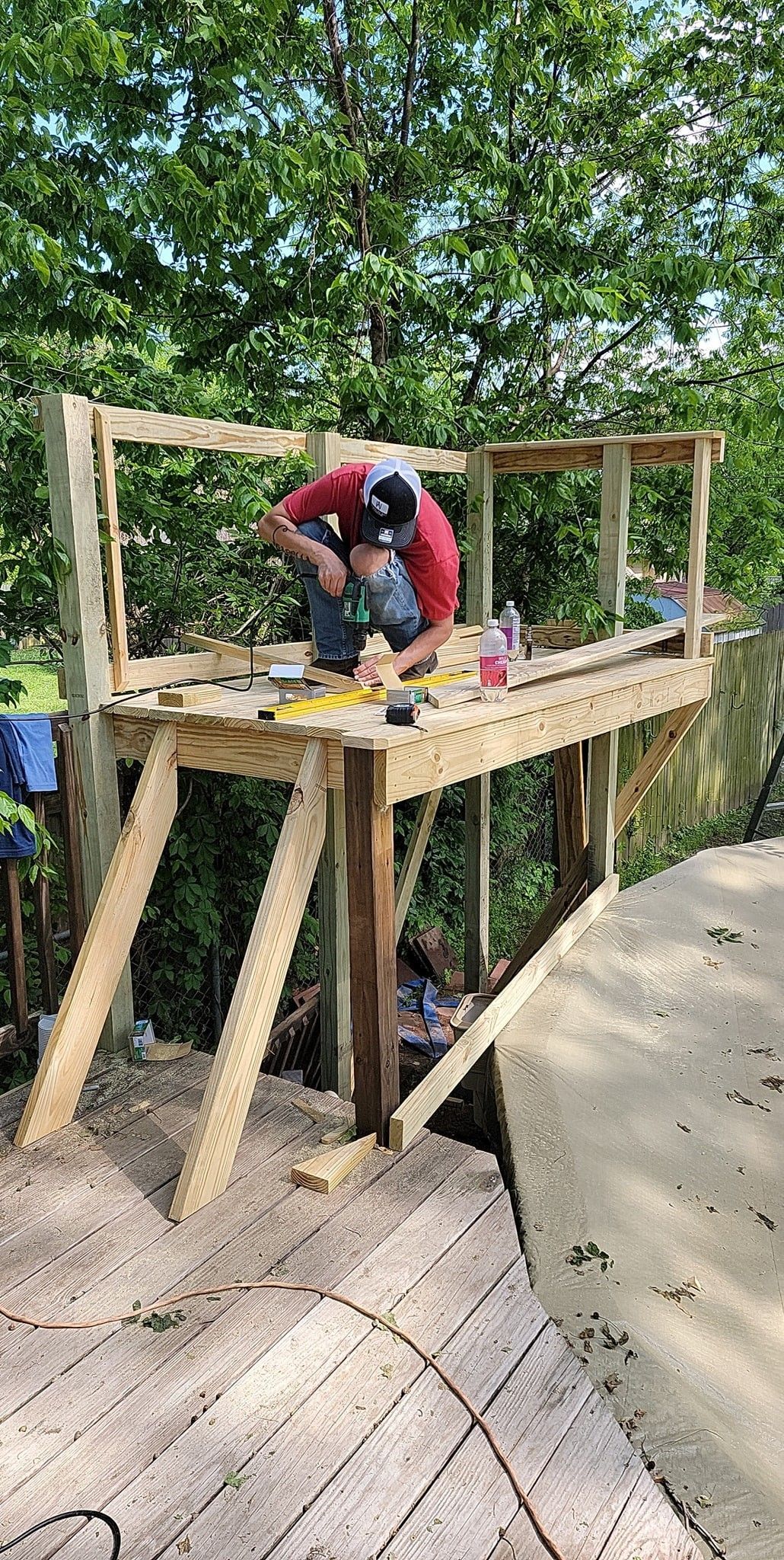 Person building a wooden deck platform in a wooded backyard, using tools and leaning over the frame.