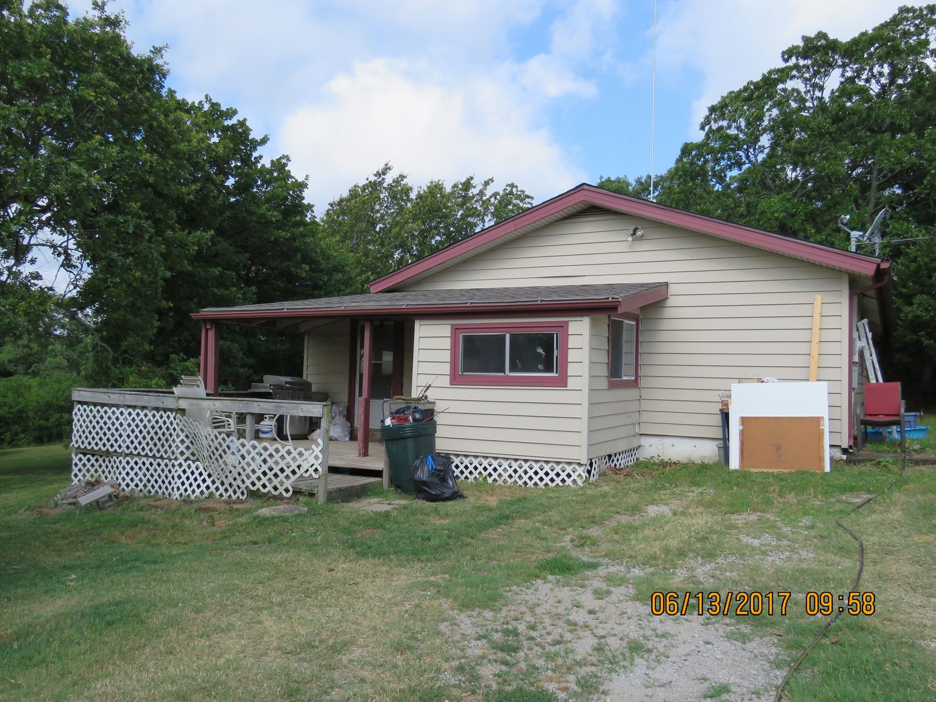 Small beige house with red trim, porch, and a grassy yard surrounded by trees.