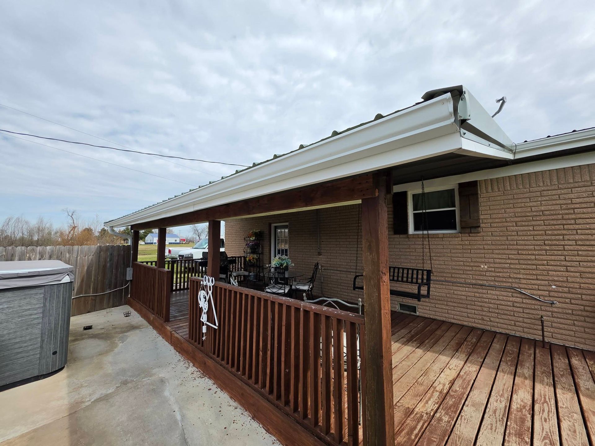 Covered wooden deck beside brick house with railing, patio furniture, and above-ground pool in a backyard.