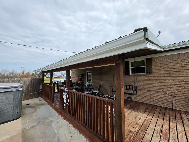 Covered wooden deck beside brick house with railing, patio furniture, and above-ground pool in a backyard.
