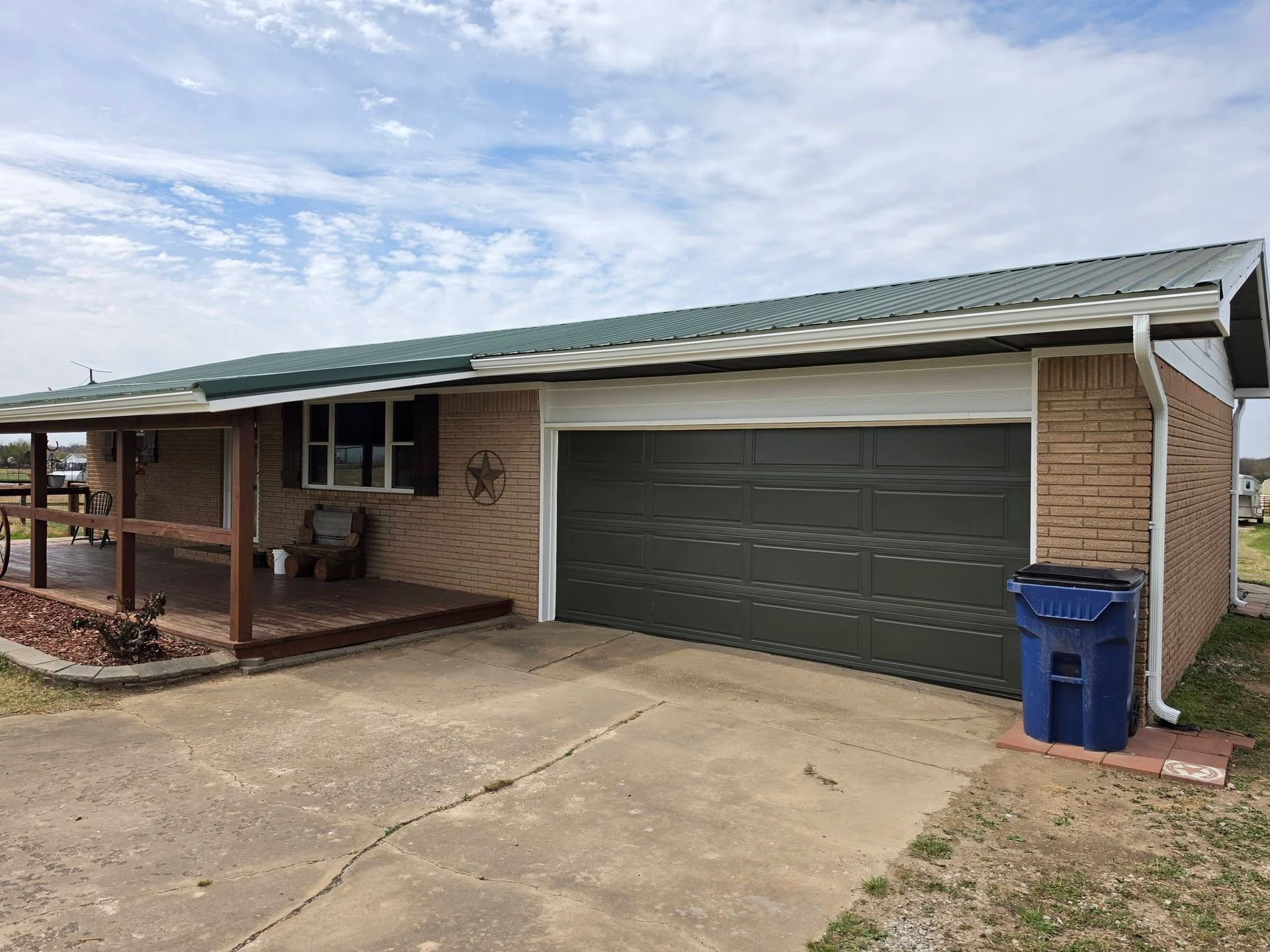 Single-story brick house with attached green garage, driveway, and blue recycling bin