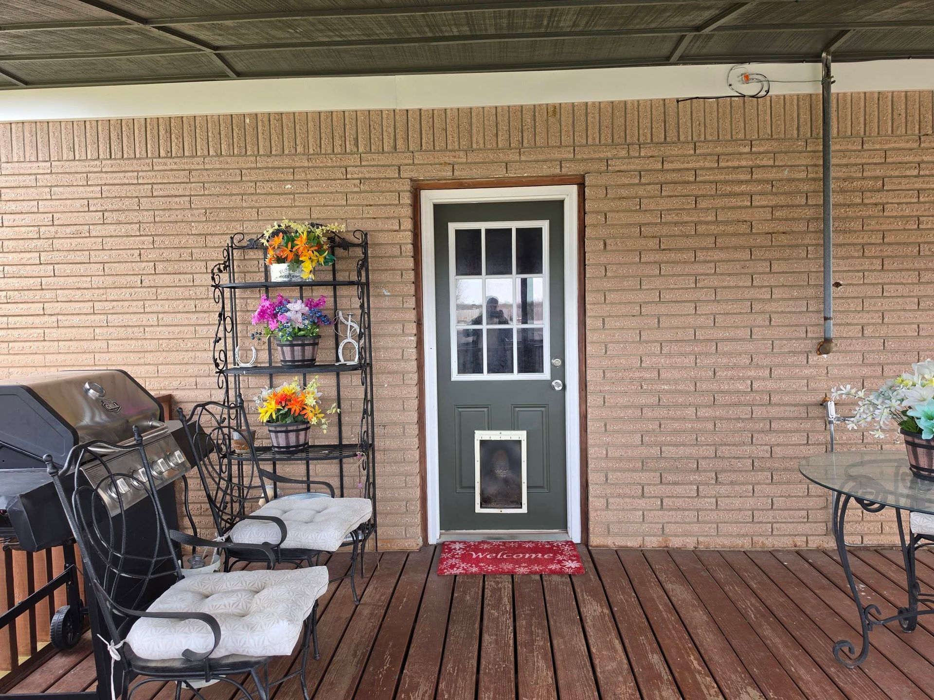 Front porch with green door, black chairs, flower stands, and red wooden decking against a brick wall