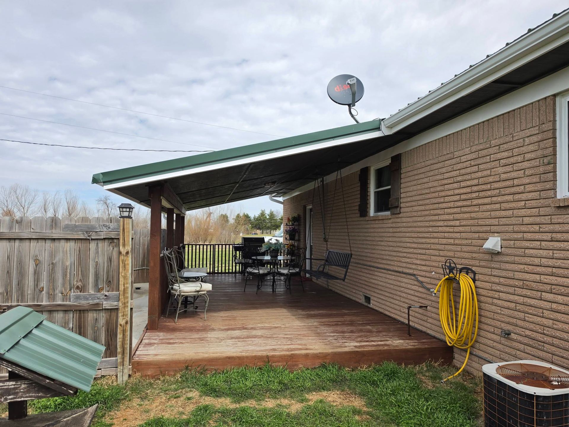 Covered backyard patio with brick wall, wooden deck, table and chairs, satellite dish, and garden hose.