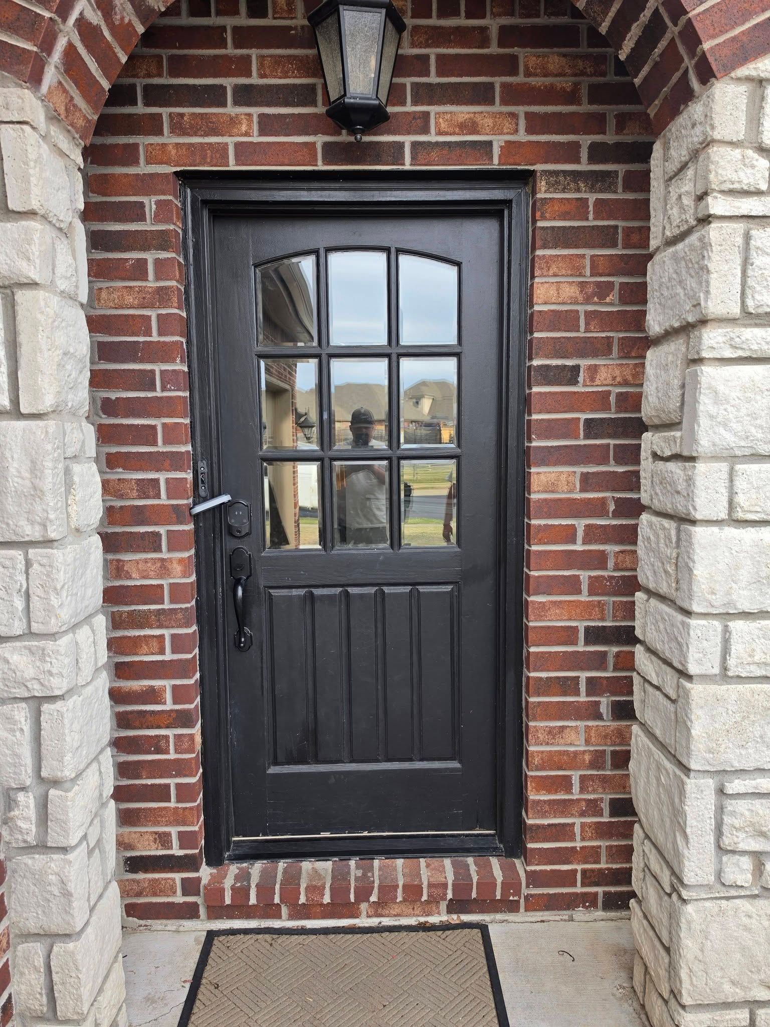 Black front door with arched window in a brick house entryway, under a wall lantern.