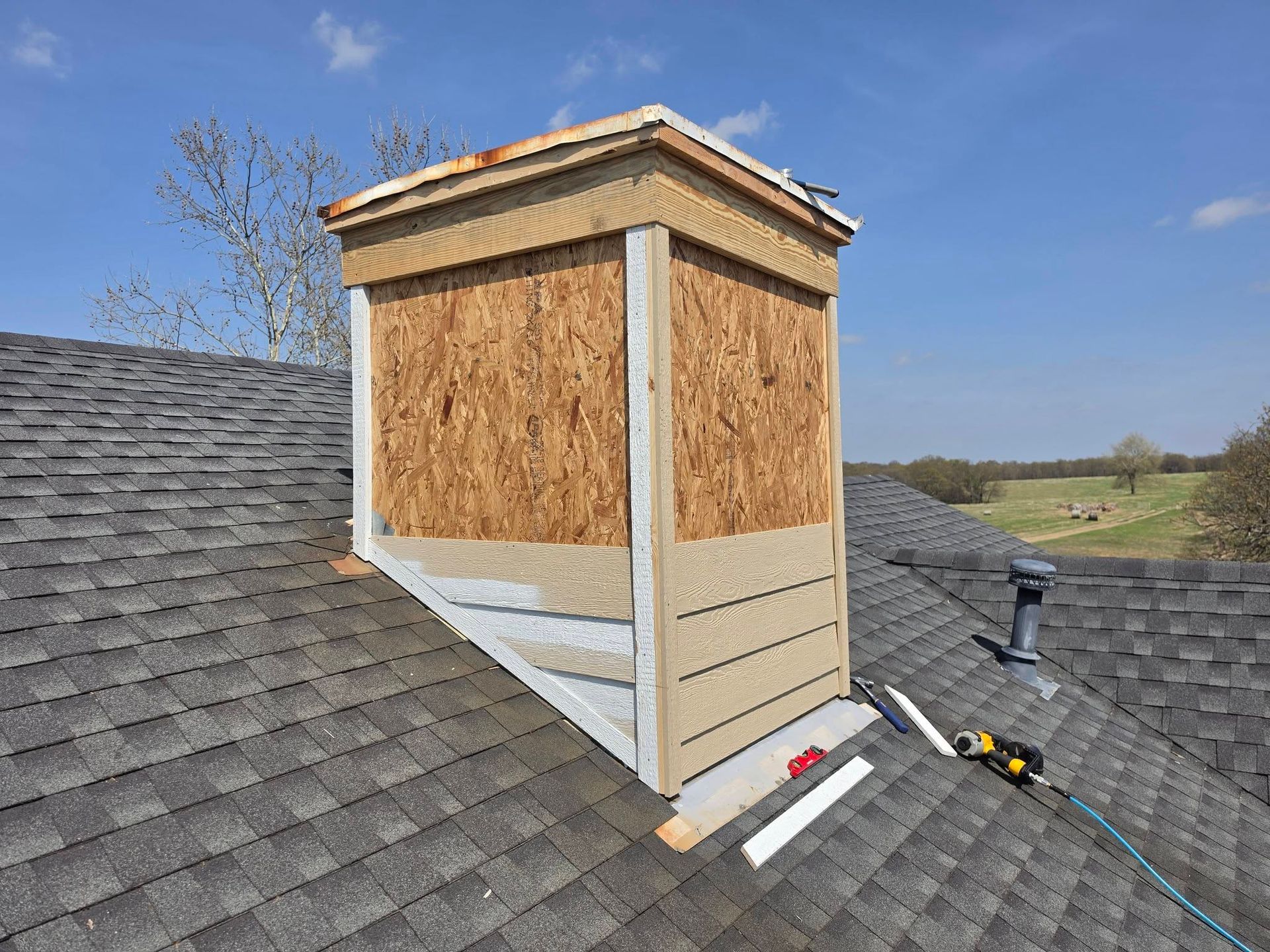 Roof chimney under construction with plywood siding and shingles on a house rooftop.