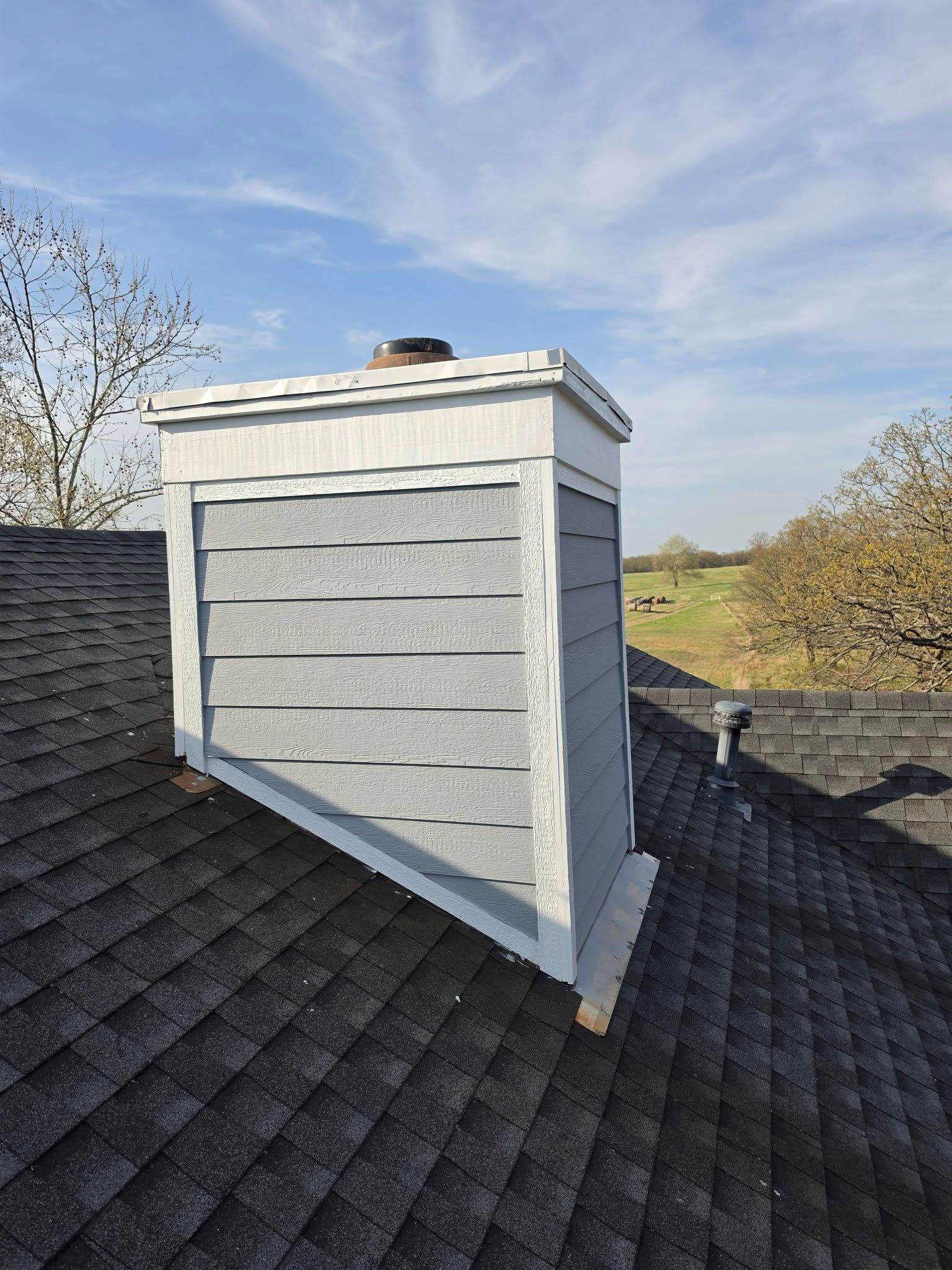 Gray rooftop chimney on a shingled roof under a blue sky.
