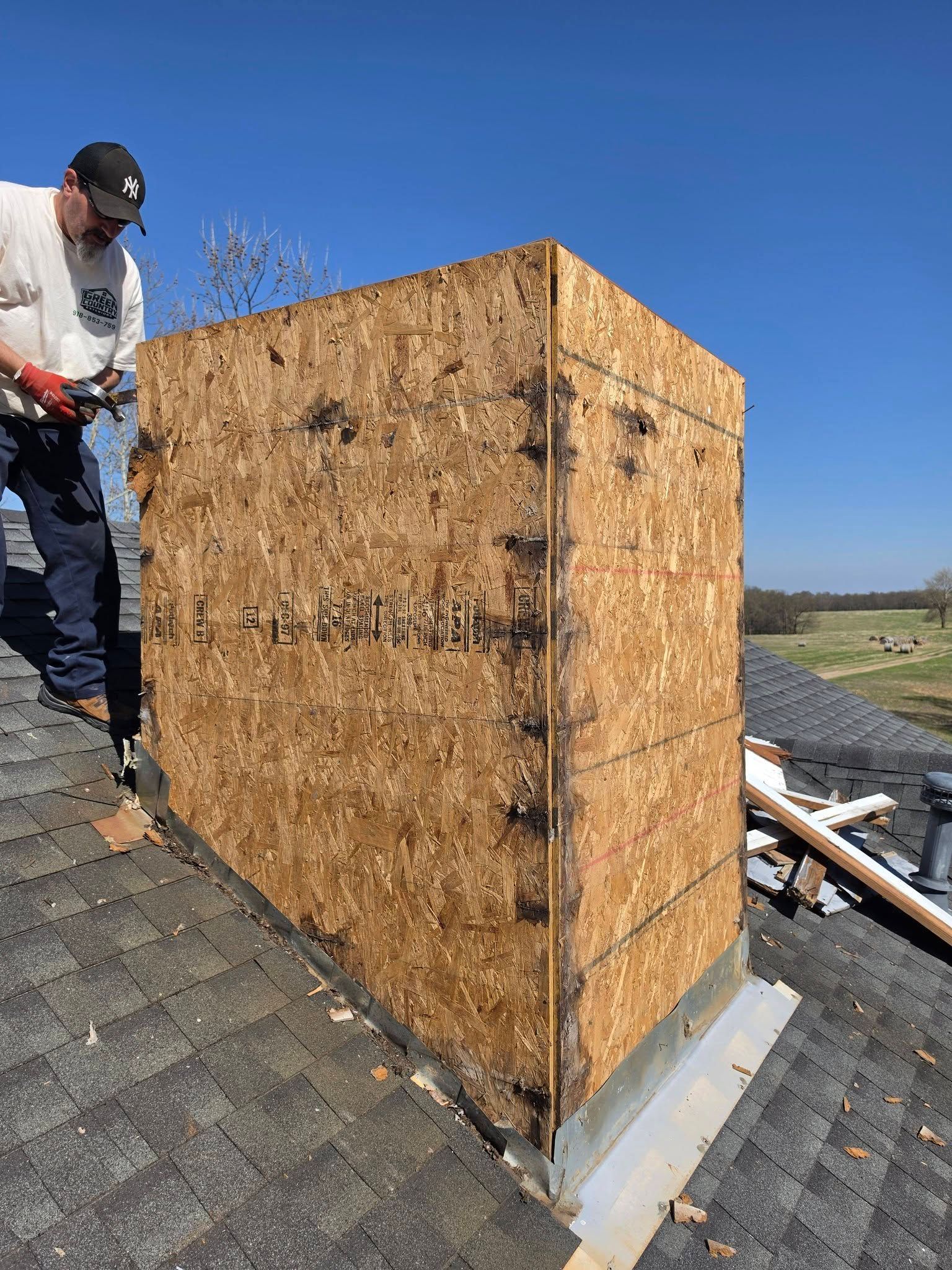 Worker on a shingled roof installing a plywood chimney chase under a clear blue sky